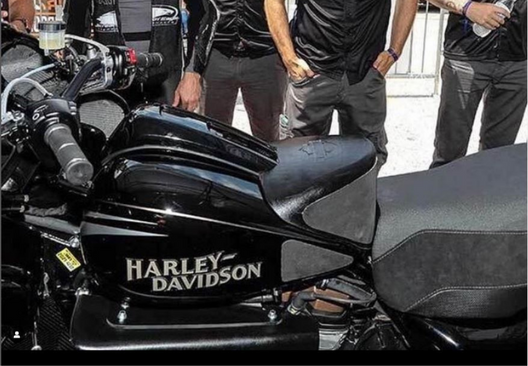 3D printed ergonomic tank extender fitted to a harley-davidson motorbike. Five bikers are stood in the background.