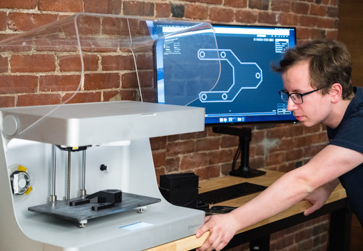 A man inspecting a 3D printer. A CAD design is open on the computer next to it.