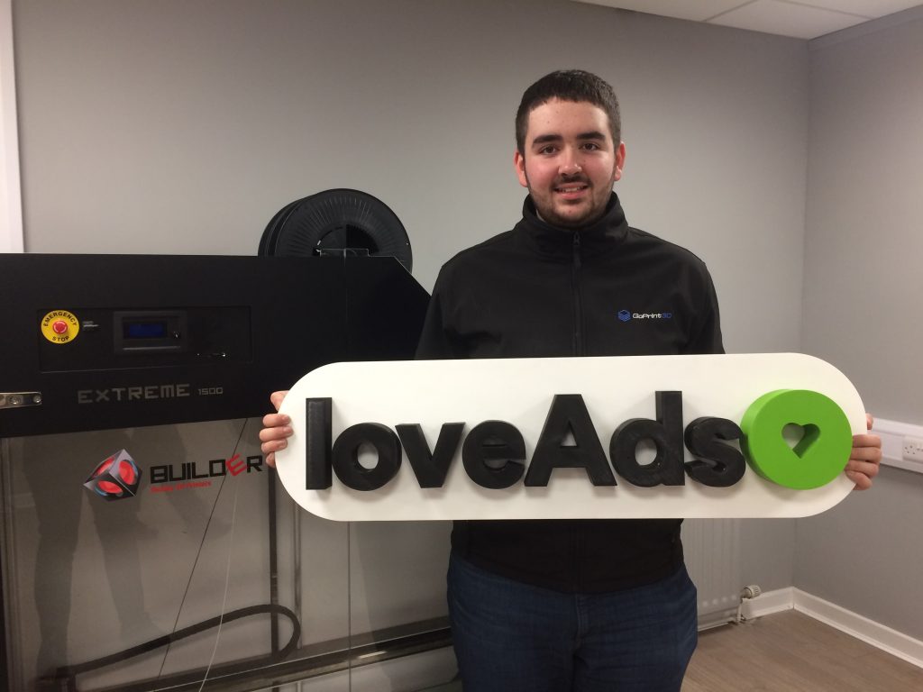 large 3d printed "love ads" logo being held by an additive x employee in front of the builder extreme 1500, the machine that printed the part