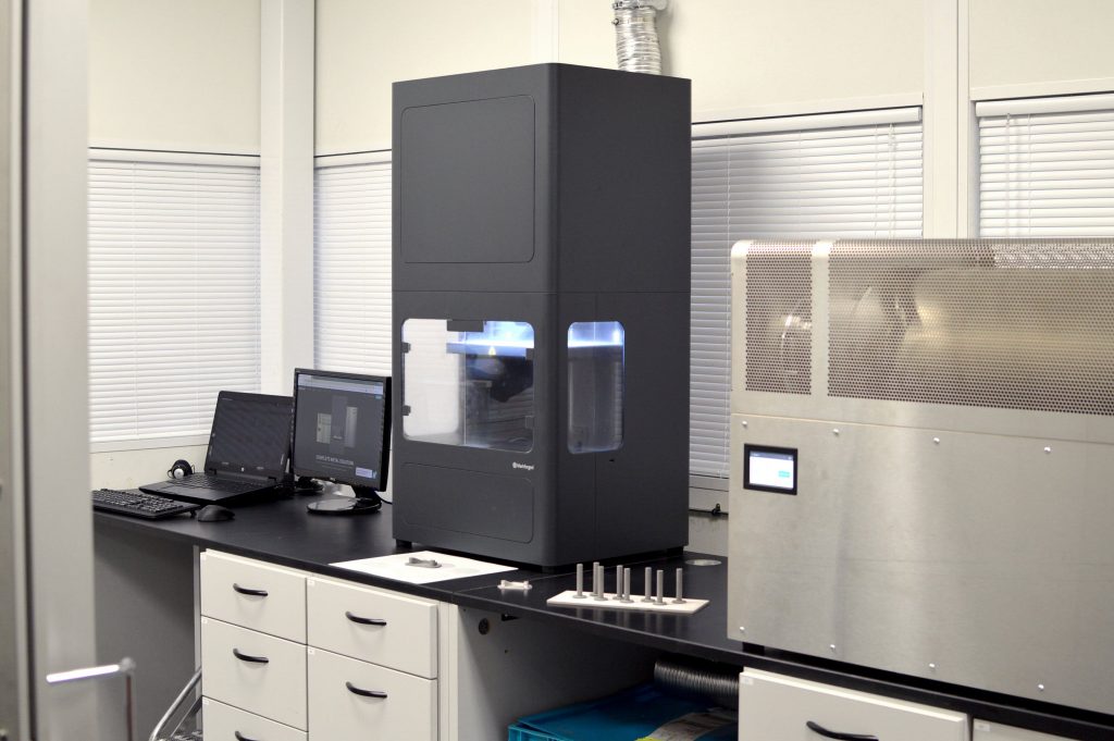 a metal x7 printer sat on a desktop along with laptops and a computer