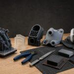 3D printed parts and post-processing tools including sanding blocks, files, cutters and safety equipment arranged on a workbench against a dark background.