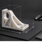 3D-printed bracket measured within a dimensional wireframe on a gridded build plate, with a digital calliper and ruler against a dark grey background, illustrating 3D printer build volume and print size.