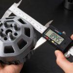 Digital callipers measuring a 3D-printed mechanical component to check dimensional accuracy and 3D printing tolerances in a dark workshop.