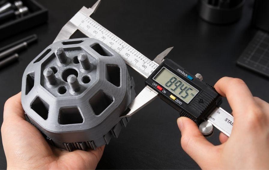 Digital callipers measuring a 3D-printed mechanical component to check dimensional accuracy and 3D printing tolerances in a dark workshop.