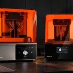Two Formlabs 3D resin printers, Form 4 and Form 3+, displaying resin-printed parts inside their orange enclosures on a workbench in a dark, industrial workshop.